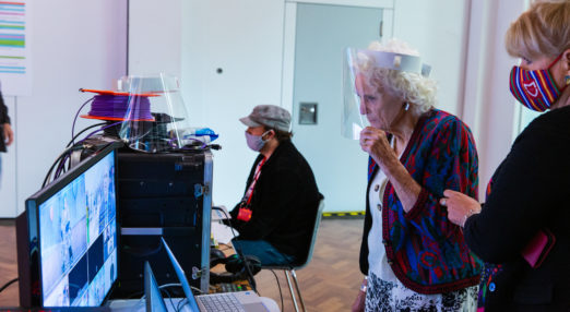 Lady Irene Morris wearing a viser watching a discussion on the studio monitor in the foreground with a member of the tech team wearing a facemask sat at his monitor.