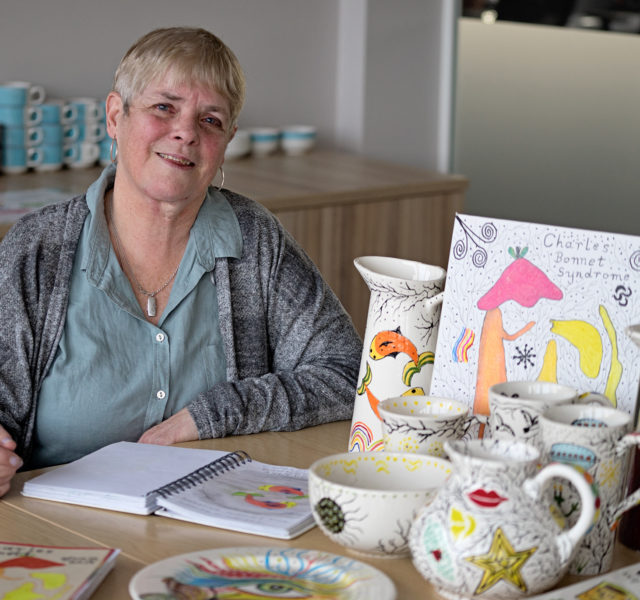 Artist Joan sitting with a display of her colourfully decorated ceramic jugs, bowls and cups and artwork. She created the pieces for a solo exhibition 'How I See My World' for Henshaws.