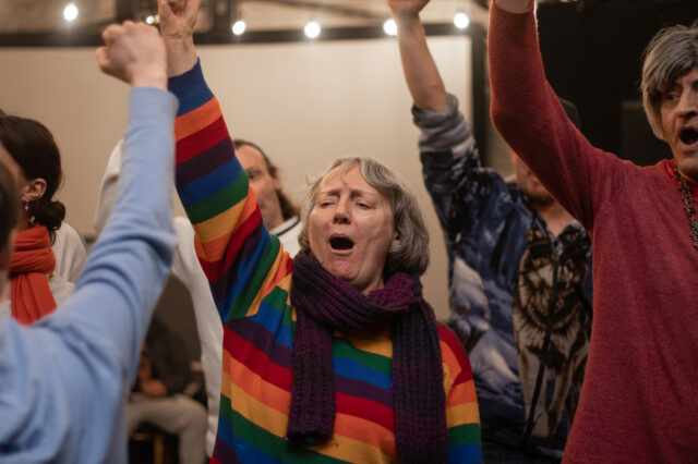 Woman in rainbow jumper at front singing with her hand in the air