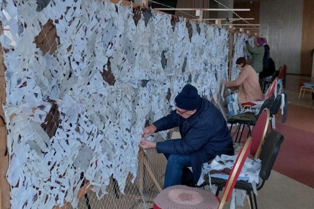 Image of creators adding symbolic lines of poetry, woven hearts and ribbons to a camouflage net.