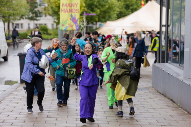Image of a parade - a crowd of people in bright clothes walking down oxford raod in Manchester. The many hands parade by Ruth Jones.