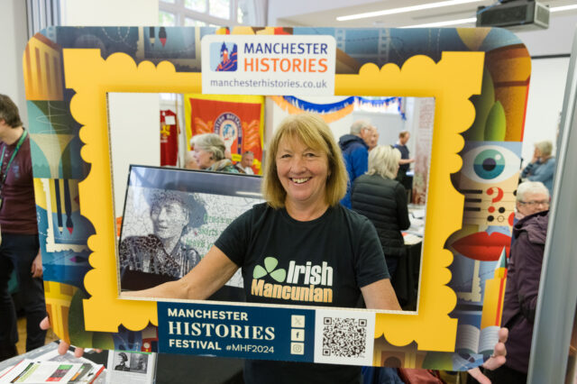 Woman holding up a Manchester Histories Frame - The woman is wearing a Irish Mancuian T-shirt and her history stall and visitors are in the background.