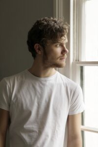 Image of Tom coly - young man dressed in white T-shirt looking out of a window