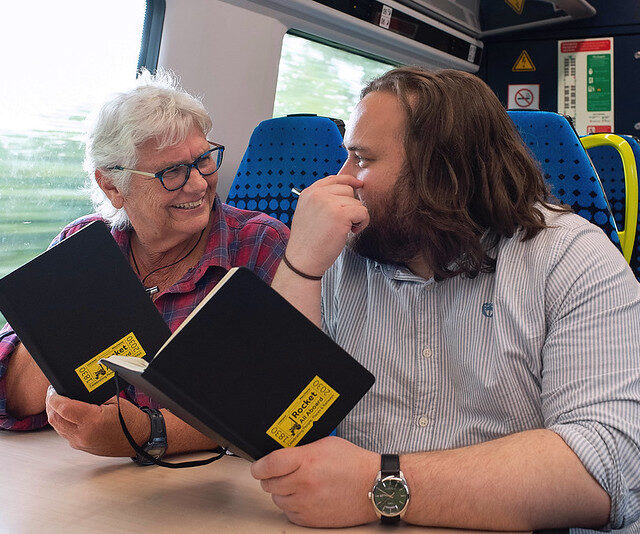 Two people sat on a moving train chatting to get each - each is holding a black book.