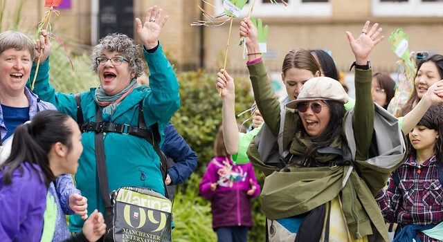 A group of people of different ages joyfully walking in an outdoor parade, smiling, cheering and waving colourful ribbon wands in a city square.