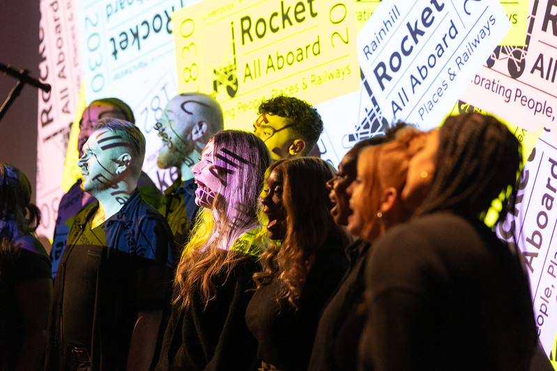 A choirs of people singing with Rocket: All Aboard logo projected onto their faces