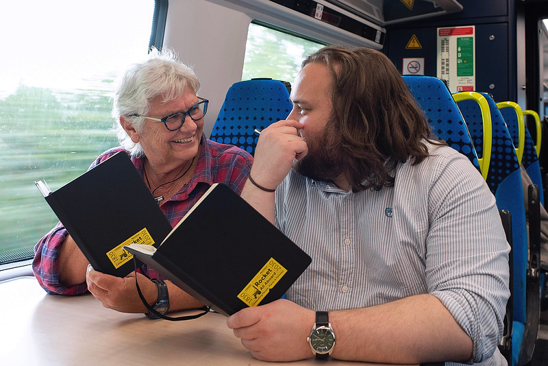 A male and female sat on a train with books in their hand talking about poetry