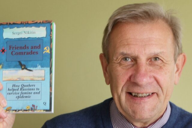 Sergei Nikitin wearing a blue V-neck jumper over a collared shirt, smiling, looking directly into the camera and holding his book 'Friends and Comrades'.