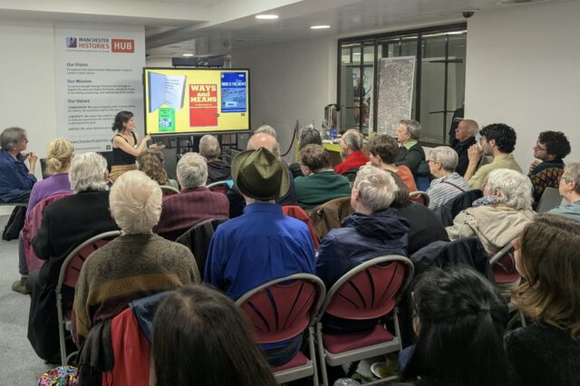 A seated audience listens to a speaker presenting in the Manchester Histories Hub, with slides displayed on a large screen and a Manchester Histories Hub logo and text on the wall behind the speaker.