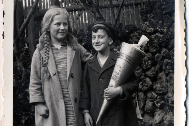 Black-and-white vintage photo of two children standing outdoors. A girl with long braids and a coat stands beside a smiling boy holding a large cone-shaped package; a wooden fence and bushes are behind them.