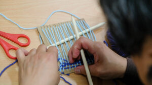 Image of a hand loom with a persons hands weaving thread through the loom