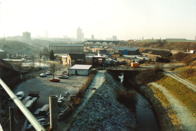 Elevated view of an industrial area with warehouses and parked cars beside a narrow canal or river, with a hazy city skyline and arched railway viaduct in the distance.