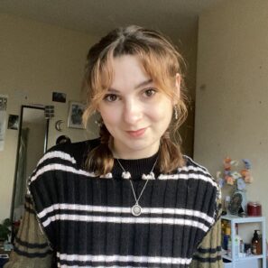 A person with auburn hair in pigtails and hoop earrings smiles at the camera indoors, wearing a black striped knit jumper and layered necklaces, with shelves in the background.