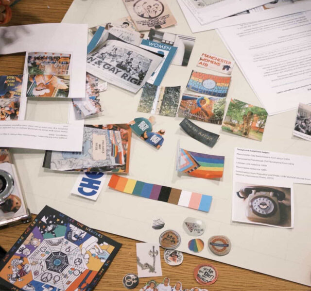 A cluttered table covered with papers, photos, pamphlets, and colorful badges, including LGBTQ+ pride materials and historical references, arranged for a preliminary design for the tapestry panel..