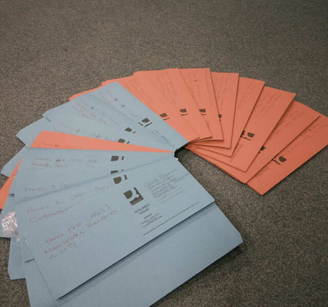 Fan of blue and orange archival document envelopes spread on a carpeted floor, each labeled with handwritten notes about which research theme they contain.