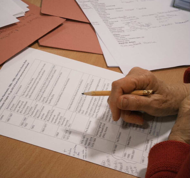 A cluttered table with folders of research notes and a researcher's hand holding a pencil making notes.