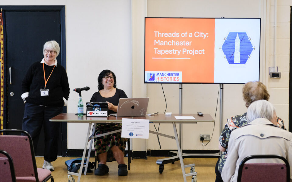 Lisa Gold leads a community talk about the Threads of a City: Manchester Tapestry Project. A member of the Textile Society stands smiling beside a table while Lisa sits with a laptop and microphone. A large screen displays the project title and the Manchester Histories logo, and audience members are seated facing the presentation in a bright community hall.
