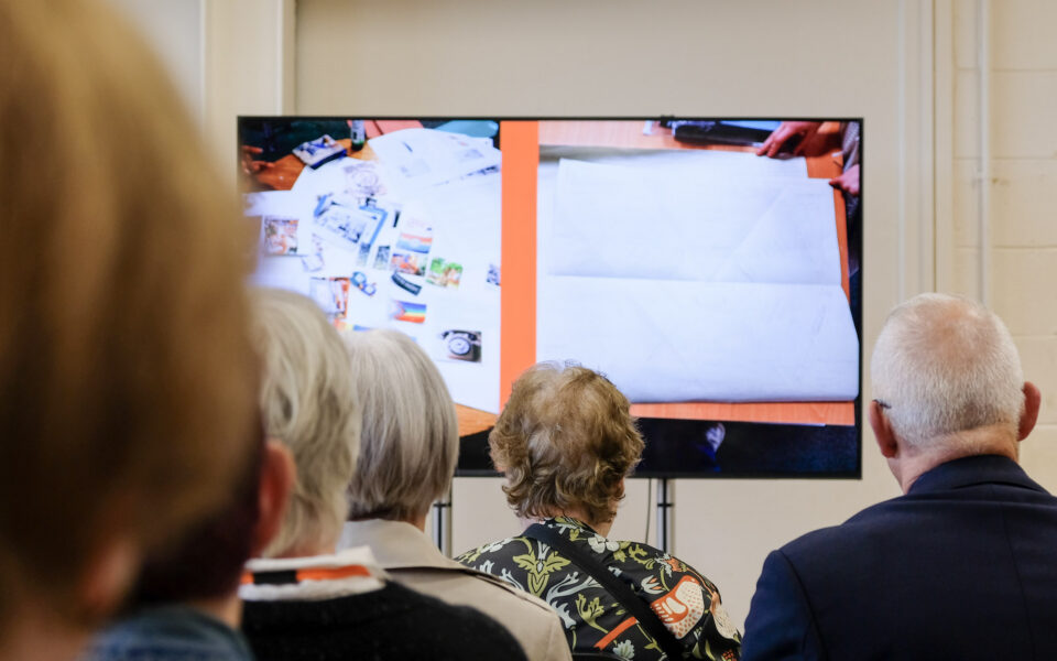 Audience members sit facing a large presentation screen showing images from the Threads of a City: Manchester Tapestry Project. The slide displays photographs of early design sketches and planning materials, while the backs of attendees' heads are visible in the foreground as they watch the presentation.
