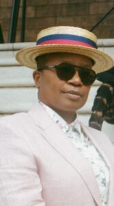 Person wearing a light pink blazer, floral shirt, straw boater hat with a red and blue ribbon, and dark sunglasses, standing outdoors on stone steps.