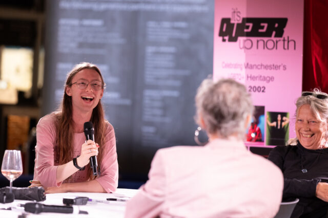 A person with long hair and glasses speaks into a handheld microphone during a Queer Up North discussion, while another participant smiles beside them.