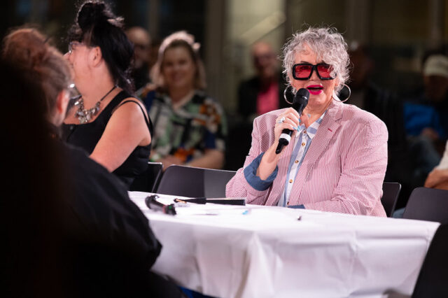 A person in a pink striped jacket and red sunglasses speaks into a handheld microphone during a group discussion.