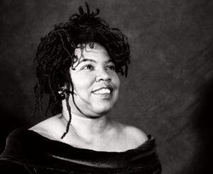 Black-and-white portrait of Cheryl Martin smiling and looking upwards, wearing an off-the-shoulder black top.