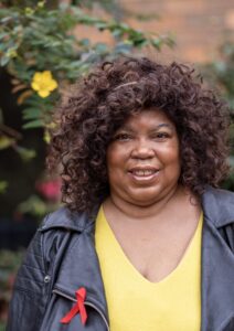 Portrait of Cheryl Martin wearing a yellow top and black leather jacket with a red ribbon pinned to it, standing outdoors.