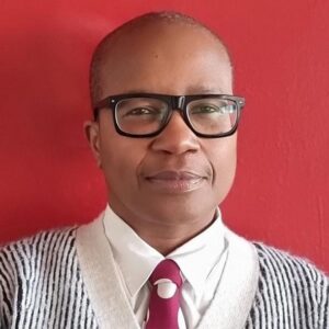 Portrait of Claud Cunningham wearing black glasses, a white shirt, patterned tie and striped cardigan against a red background.