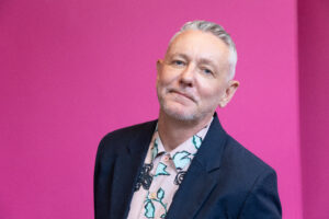 Portrait of a man wearing a dark blazer and patterned shirt against a bright pink background.