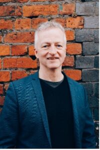 Portrait of John McGrath standing in front of a red brick wall, wearing a dark blue jacket and black top.