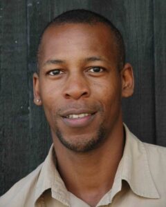 Portrait of Topher Campbell looking at the camera, wearing a light-coloured shirt against a dark wooden background.