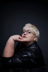 Portrait of a person with short platinum-blonde hair and round glasses, wearing a black leather outfit against a dark background.