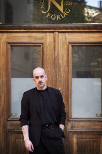 Portrait of Andrew McMillan standing in front of wooden doors, wearing a black suit and looking towards the camera.