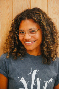 Portrait of Ella Otomewo smiling at the camera, with curly hair and glasses, standing in front of a wooden background.