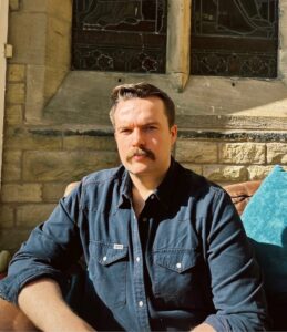 Portrait of Louis Glazzard seated indoors in sunlight, wearing a dark blue shirt and looking towards the camera.
