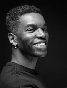 Black-and-white portrait of Okechukwu Nzelu smiling towards the camera, wearing a dark top and a single drop earring.